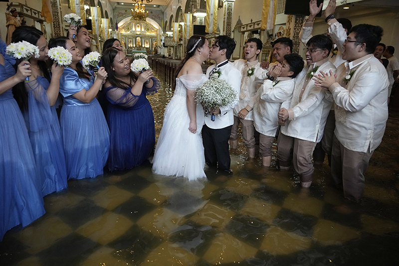 Wedding in the Flood by Aaron Favila _ Credit: © Aaron Favila, Associated Press. Title: Wedding in the FloodCredit: © Aaron Favila, Associated PressCaption: The newlyweds share a kiss as guests cheer. The couple have been together for ten years. According to Verdillo, “This is just one of the struggles that we’ve overcome.” Malolos, Bulacan province, Philippines, 22 July 2025.Story: When Typhoon Wipha hit the Philippines and flooded Barasoain Church, Jade Rick Verdillo and Jamaica Aguilar faced a difficult decision: should they cancel their wedding or proceed with the marriage? The couple carried on despite high waters, a testament to love and resilience in the face of severe weather. Located on a delta, Bulacan province is vulnerable to more frequent and extreme floods caused by aging drainage systems, dredging projects, overextraction of groundwater, and climate change.